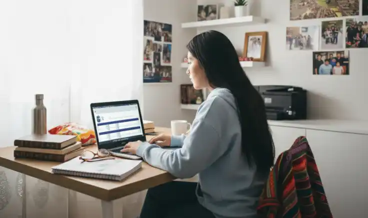 Joven estudiando contabilidad y finanzas a distancia en su laptop desde casa, rodeada de libros, apuntes y una taza de café en un espacio de estudio acogedor.
