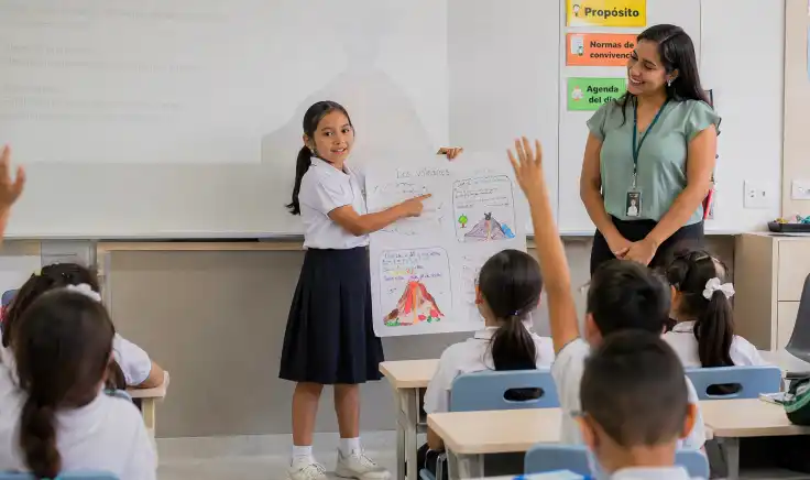Una maestra de primaria sonríe y apoya a una alumna que muestra un dibujo a sus compañeros de clase, promoviendo el desarrollo de la expresión oral y la confianza en la escuela.
